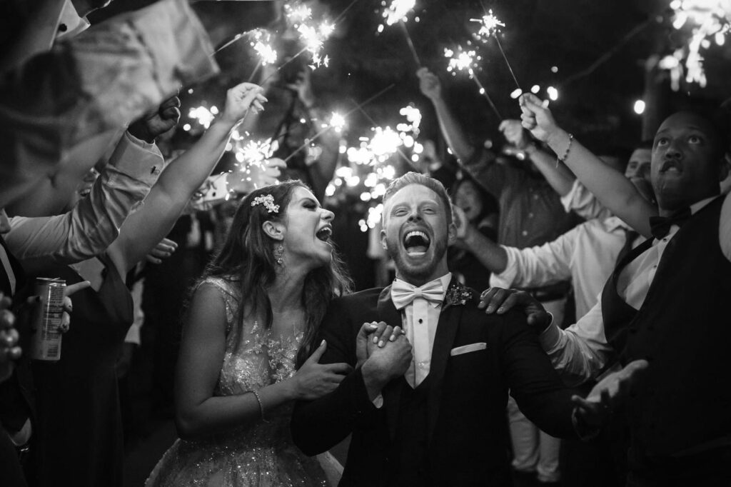 A bride and groom laugh while exiting their wedding under sparklers.